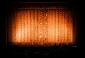 A view of a theatre stage in a darkened auditorium, with rust-colored curtains lit by spotlights.